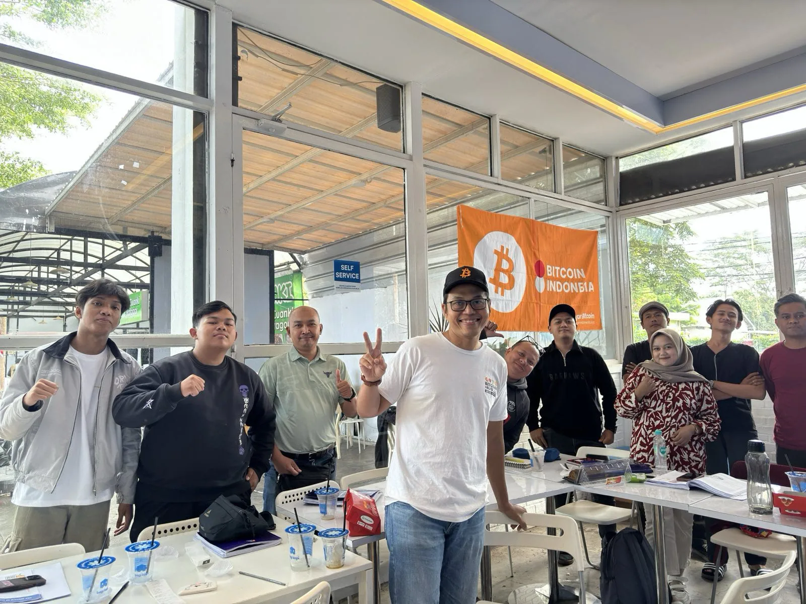 My First Bitcoin instructor with students in front of a Bitcoin Indonesia banner, giving a peace sign