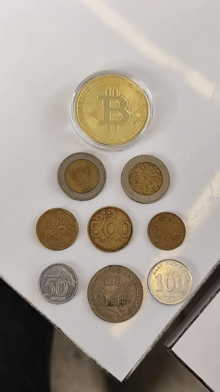 A gold-plated Bitcoin coin next to Indonesian Rupiah coins on a table