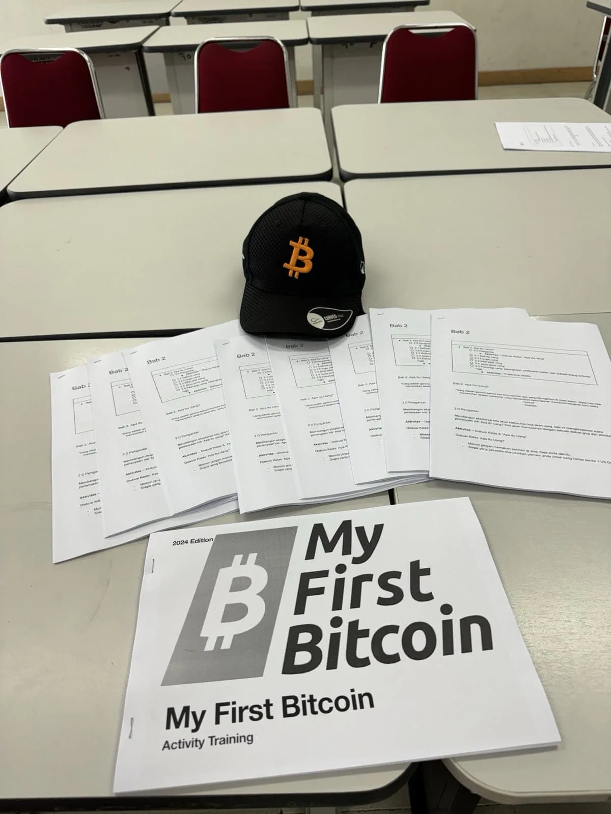 Stack of My First Bitcoin activity training workbooks and a Bitcoin cap on classroom desks