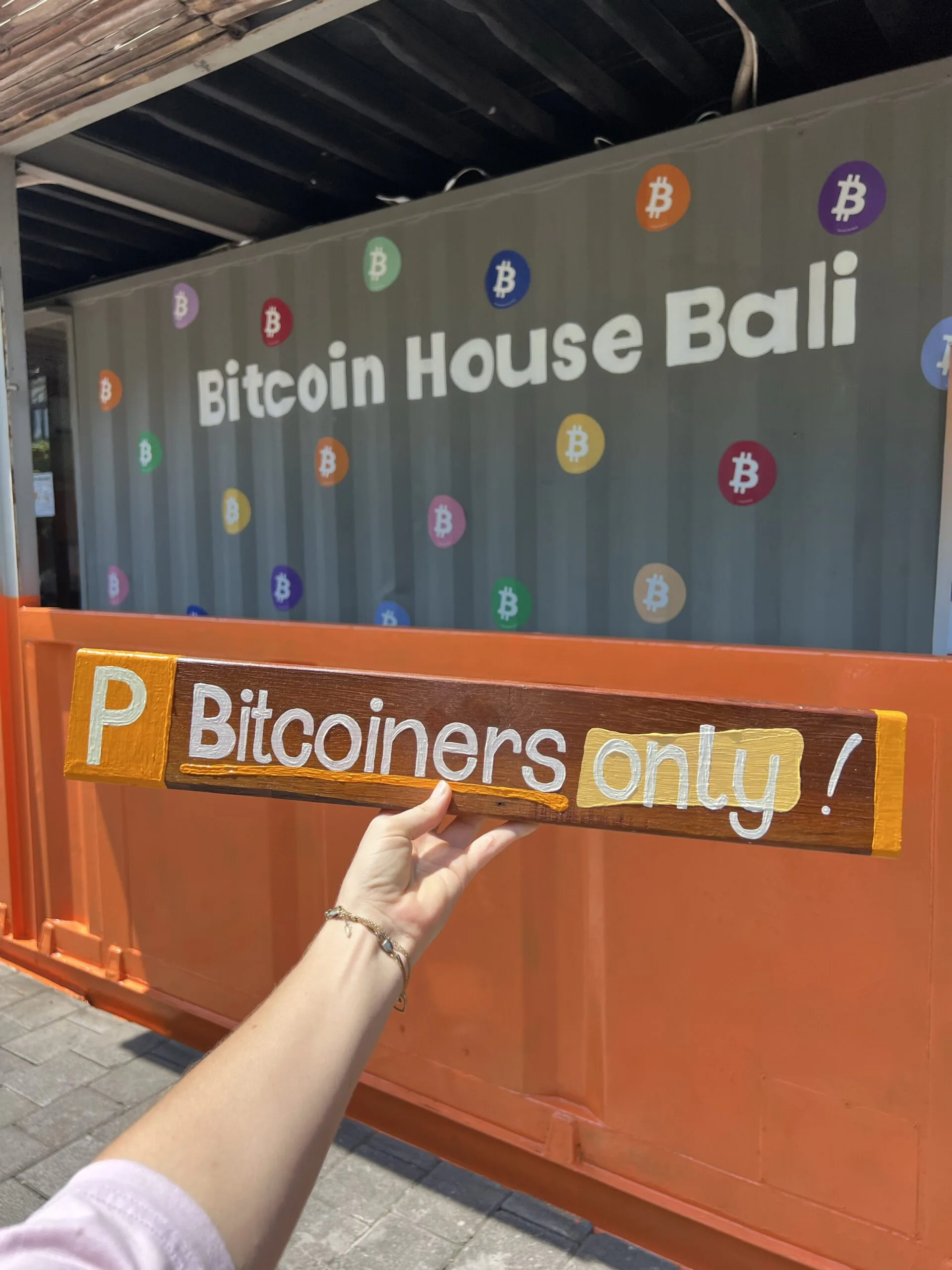 Bitcoin House Bali shipping-container building covered in colorful Bitcoin logo stickers, with a hand-painted Bitcoiners Only parking sign