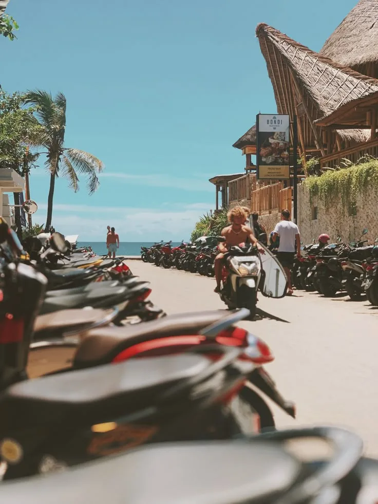 A street in Canggu, Bali lined with parked motorbikes, a surfer riding past toward the beach